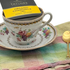 A beautiful white teacup and saucer with a floral pattern. The teacup has a tea bag inside. The teacup and saucer are on a multi-colored checkered napkin with a gold teaspoon.