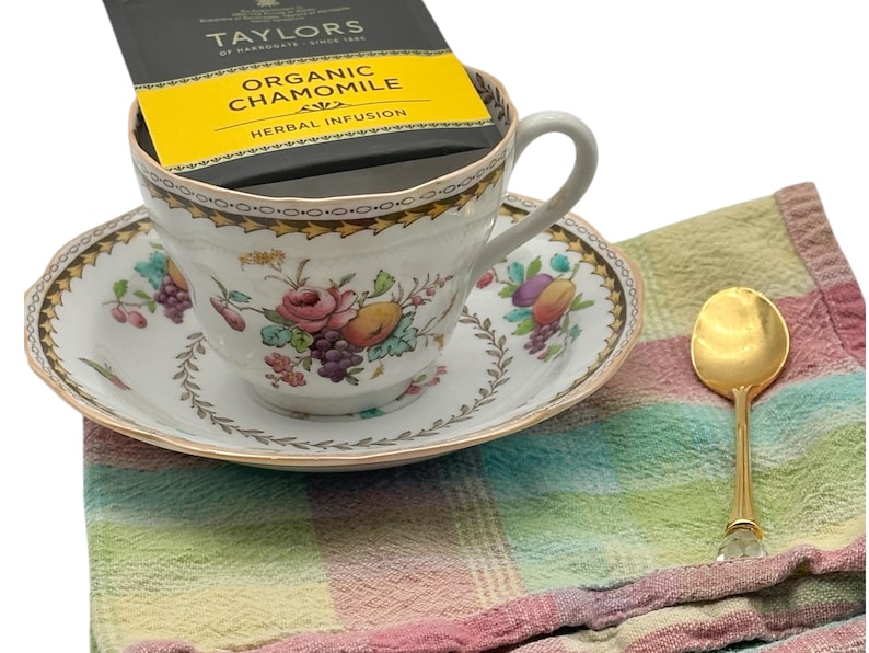 A beautiful white teacup and saucer with a floral pattern. The teacup has a tea bag inside. The teacup and saucer are on a multi-colored checkered napkin with a gold teaspoon.
