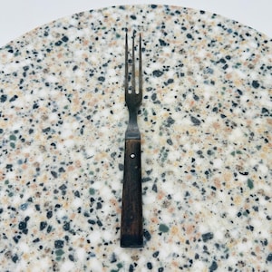 May include: An antique fork with three tines and a dark wooden handle. The metal fork shows signs of age and wear. The handle is attached with a single rivet. The fork is set against a speckled, light-colored surface.