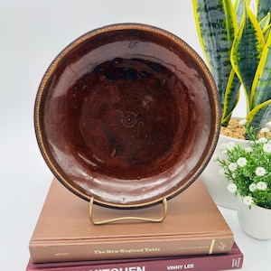 May include: A round ceramic plate with a deep, glossy, dark brown glaze and a textured, ridged rim. The plate is displayed upright on a gold-toned metal stand, showcasing a smooth, reflective surface with subtle variations in the brown color tones.