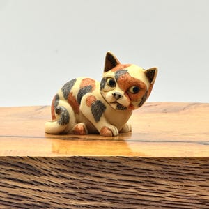 May include: A small, detailed figurine of a seated cat. The cat has a cream-colored base with patches of orange, black, and brown. The cat is sitting on a wooden surface with a light background.