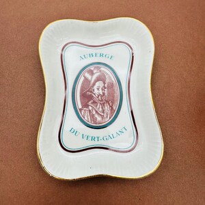May include: A white ceramic rectangular tray with scalloped edges and a gold-trimmed rim. The center features a maroon oval illustration of a man in a historical hat, surrounded by the teal text AUBERGE DU VERT-GALANT in a decorative frame.