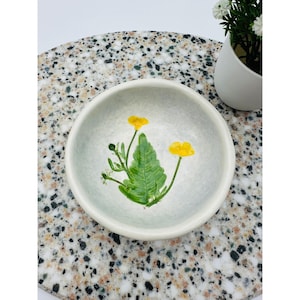 May include: A small, off-white ceramic bowl with a hand-painted floral design. The design features two yellow flowers and a green leaf. The bowl is sitting on a speckled surface, with a small white pot of flowers in the background.