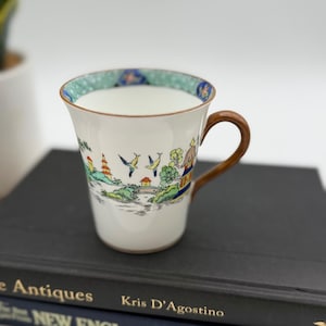 May include: A white ceramic mug with a brown handle featuring a colorful landscape illustration of pagodas, trees, and birds. The interior rim is decorated with a teal and blue patterned border. The mug has a tapered shape and a smooth finish.