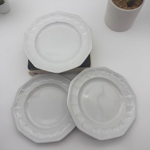 May include: Three white ceramic plates with an octagonal shape and a raised, textured rim pattern. The plates feature a smooth, glossy finish and a central circular basin. One plate is stacked on top of a dark book, while two others sit below.