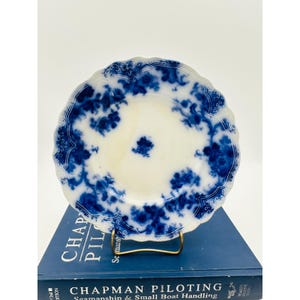 May include: A white plate with a blue floral design and a scalloped edge. The plate is displayed on a gold-colored stand, resting on a blue book with the title "CHAPMAN PILOTING Seamanship & Small Boat Handling."