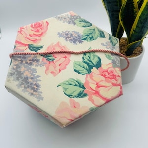 May include: A hexagonal gift box with a floral pattern featuring pink roses, purple lilacs, and green leaves on an off-white background. A pink cord wraps around the box. A potted plant is in the background.