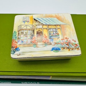 May include: A square, ceramic trinket box with a painted scene of a flower shop. The shop has a yellow facade, a blue striped awning, and is surrounded by potted flowers in shades of red, pink, and blue. The text "The Post Shop" is visible.
