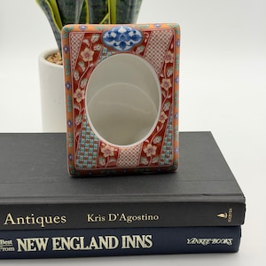 May include: A rectangular ceramic picture frame featuring a vibrant orange-red background with intricate floral patterns, blue and white geometric accents, and a central oval opening. The frame is decorated with small white flowers and green leaves, with a blue and white floral motif at the top center.