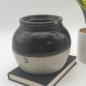 May include: A round ceramic crock featuring a two-tone design with a dark brown glazed upper section and a light beige lower section. The lower portion displays a blue number 3 printed on the front. The rim shows a small chip in the glaze.
