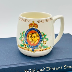 May include: Cream-colored ceramic mug featuring a royal crest with portraits, a crown, and the text "GEORGE VI & QUEEN EL". The mug has a curved shape with a handle and is set against a blue book.
