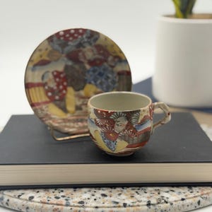 May include: A ceramic teacup and matching saucer set featuring intricate hand-painted figures in red, blue, and yellow attire. The cup has a small handle and a light-colored interior, while the saucer displays a detailed scene with patterned clothing and gold accents throughout the design.