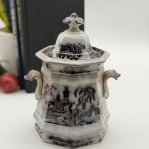 May include: An octagonal ceramic sugar bowl with a lid, featuring a black and white transferware landscape pattern of trees and buildings. The vessel has two curved side handles and a decorative finial on the lid, with a glossy finish.