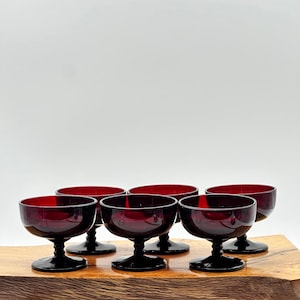 May include: Six vintage ruby red coupe glasses with black stems are arranged on a wooden surface. The glasses have a rounded bowl shape and a short stem. The background is a plain white.
