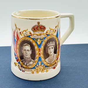 May include: Cream-colored ceramic mug with gold trim, featuring portraits of King George VI and Queen Elizabeth, with "Coronation May 1937" text. The mug has a rectangular handle and is decorated with the Union Jack flag.