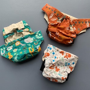 Könnte beinhalten: Drei wiederverwendbare Stoffwindeln mit verschiedenen Tiermustern. Eine Windel ist grün mit weißem Hintergrund, eine andere ist orange mit weißem Hintergrund und die letzte ist weiß mit braunem Hintergrund.
