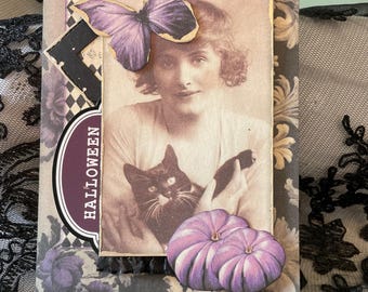 May include: A Halloween-themed greeting card featuring a vintage sepia-toned image of a woman holding a black cat. The card is adorned with a purple butterfly, pumpkins, and the word "HALLOWEEN". The background has a floral pattern.