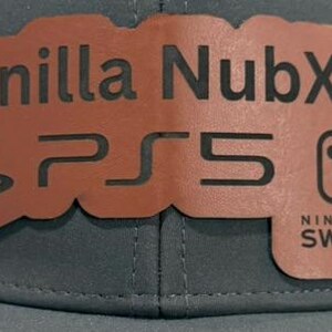 May include: A brown leather patch with the text "Vanilla NubXX" and the logos for PlayStation 5 and Nintendo Switch. The patch is attached to a gray baseball cap.