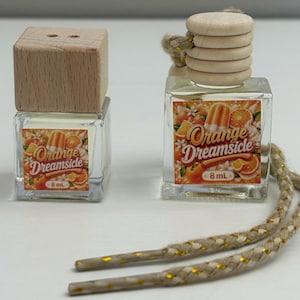 May include: Two small, clear glass bottles with wooden tops and decorative labels. The labels feature the text "Orange Dreamsicle" with orange and white graphics. Each bottle has a decorative string attached.