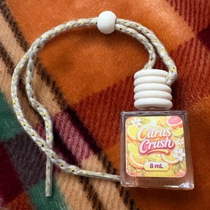 May include: A small, clear glass bottle with a white wooden top and a decorative label that reads "Citrus Crush" with images of citrus fruits. The bottle is attached to a beige and gold rope, and contains 8 mL of liquid.