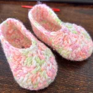 Op de afbeelding: Een paar gehaakte babyschoentjes in roze, wit en groen. De schoentjes zijn gemaakt van zacht, getextureerd garen en hebben een eenvoudig, klassiek ontwerp.