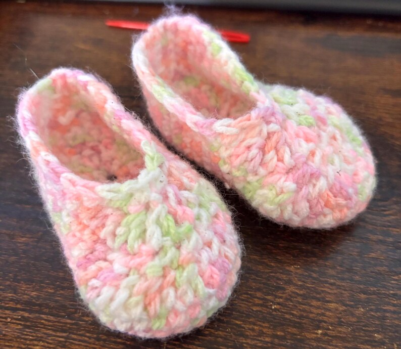 Op de afbeelding: Een paar gehaakte babyschoentjes in roze, wit en groen. De schoentjes zijn gemaakt van zacht, getextureerd garen en hebben een eenvoudig, klassiek ontwerp.
