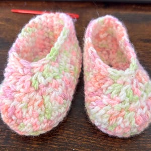 Op de afbeelding: Een paar gehaakte babyschoentjes in roze, groen en wit. De schoentjes hebben een simpel ontwerp en zijn gemaakt van zacht garen.