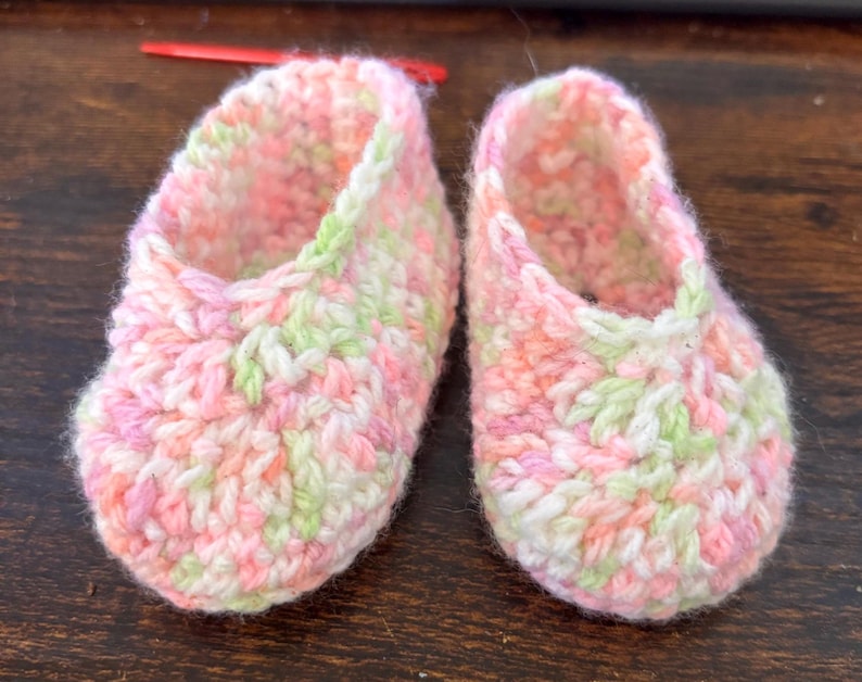 Op de afbeelding: Een paar gehaakte babyschoentjes in roze, groen en wit. De schoentjes hebben een simpel ontwerp en zijn gemaakt van zacht garen.