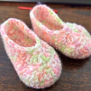 Op de afbeelding: Een paar roze, groene en witte gehaakte babyschoentjes. De schoentjes zijn gemaakt van een zacht, getextureerd garen en hebben een eenvoudig, klassiek ontwerp.