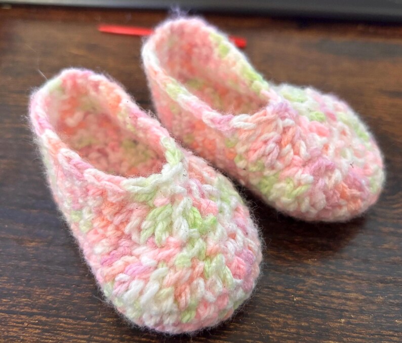 Op de afbeelding: Een paar roze, groene en witte gehaakte babyschoentjes. De schoentjes zijn gemaakt van een zacht, getextureerd garen en hebben een eenvoudig, klassiek ontwerp.