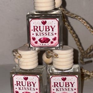 Könnte beinhalten: Drei quadratische Glasflaschen mit hellen Holzverschlüssen und Aufhängeschnüren. Jede Flasche hat ein weißes Etikett mit dem roten Schriftzug "RUBY KISSES", verziert mit Herz- und Edelsteinmotiven. Jedes Etikett zeigt deutlich das Volumen 8 mL.