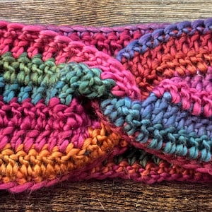 Könnte beinhalten: Ein farbenfrohes gehäkeltes Stirnband mit einem gedrehten Design. Das Stirnband hat Streifen aus rosafarbenem, orangefarbenem, grünem, blauem und violettem Garn.