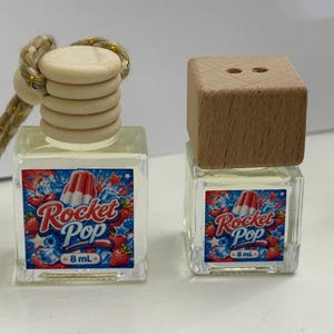 Puede incluir: Dos pequeñas botellas de vidrio transparente con tapas de madera. Cada botella tiene una etiqueta con el texto "Rocket Pop" y un gráfico de un helado tricolor. Las botellas están llenas de un líquido transparente y tienen 8 ml.