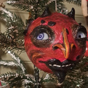 May include: A round, red, textured ornament shaped like a devil's head with two small black horns, a pointed yellow nose, a black goatee, and wide, light blue eyes. The ornament features a string loop at the top for hanging on a tree branch.