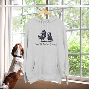 May include: A light gray hoodie with a front pocket and a drawstring hood. The hoodie features a graphic of two black birds with the text "Two Birds One Stoned" in a decorative font. The hoodie is hanging on a wooden hanger.