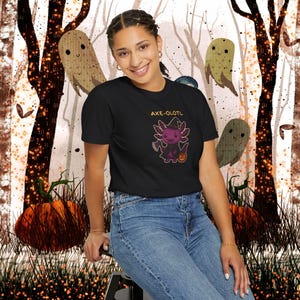 May include: Black t-shirt featuring a Halloween-themed axolotl design with the text "AXE-OLOTL". The axolotl is depicted in a witch costume holding an axe and a jack-o'-lantern. The background includes cartoon ghosts, pumpkins, and a forest scene.