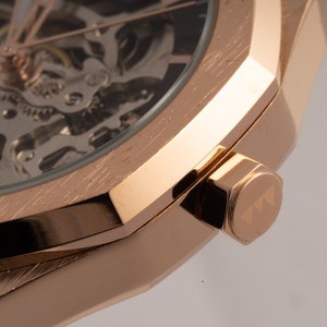May include: Close-up of a rose gold watch with a skeletonized dial and a brushed finish. The watch has a square case and a crown with the letter 'T' engraved on it.