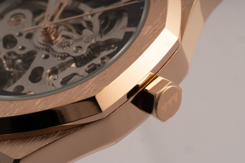 May include: Close-up of a rose gold watch with a skeletonized dial and a brushed finish. The watch has a square case and a crown with the letter 'T' engraved on it.