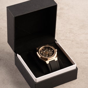May include: A gold-tone watch with a black rubber band is displayed in a black box with a white interior.