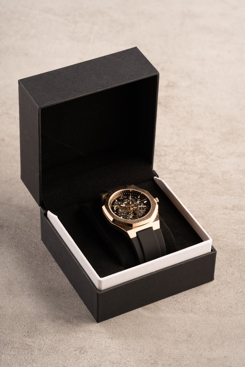 May include: A gold-tone watch with a black rubber band is displayed in a black box with a white interior.