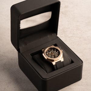 May include: A gold-tone watch with a skeleton dial and black rubber strap is displayed in a black box with a gray lining.