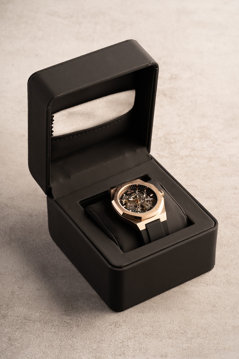 May include: A gold-tone watch with a skeleton dial and black rubber strap is displayed in a black box with a gray lining.