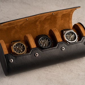 May include: A black leather watch roll with brown suede lining. The roll has four slots for watches and features silver metal snaps. Four watches with skeletonized faces are displayed in the slots.