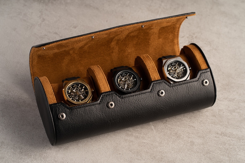 May include: A black leather watch roll with brown suede lining. The roll has four slots for watches and features silver metal snaps. Four watches with skeletonized faces are displayed in the slots.