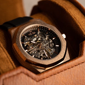 May include: A rose gold skeleton watch with a black face and a black leather band. The watch is in a brown leather watch box.