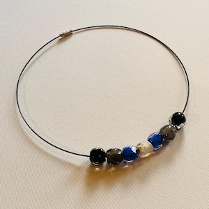 Op de afbeelding: Een zwarte draadketting met een reeks facetgeslepen kralen in zwart, grijs, blauw en helder. De kralen zijn in een gebogen lijn geregen, wat een minimalistisch ontwerp creëert. De ketting ligt op een witte ondergrond.