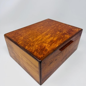 May include: A rectangular wooden box featuring a rich, swirling burl wood grain pattern on the lid and sides. The box has a smooth, polished finish with dark brown edges and a small, integrated horizontal handle on the front panel.