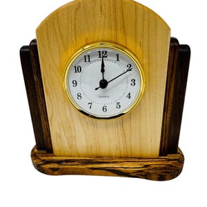 May include: A wooden quartz clock with a light-colored face and gold-colored trim. The clock has a light-colored wooden face and a dark-colored wooden base and sides. The clock hands point to 12:00. The clock is approximately 6 inches tall.