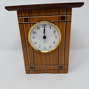 May include: A wooden mantel clock featuring a trapezoidal body with inlaid dark wood vertical and horizontal lines. The clock face is white with black Arabic numerals, a gold-toned circular bezel, and the word QUARTZ printed in black.