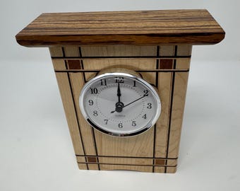 May include: A wooden clock with a light wood frame and a dark wood top. The clock face is white with black numbers and hands. The clock is sitting on a white surface.